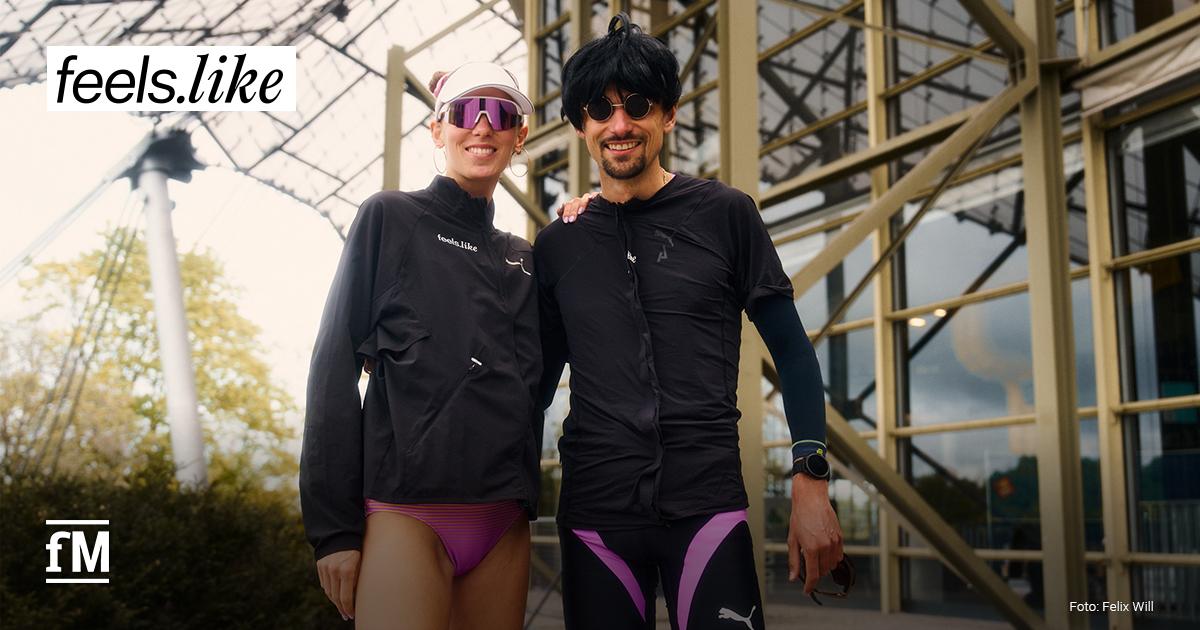 Zwei sportlich gekleidete Personen (Esther und Hendrik Pfeiffer ) stehen nebeneinander vor einer modernen Glas- und Stahlkonstruktion und lächeln in die Kamera. Beide tragen schwarze Laufkleidung mit feels.like-Branding.