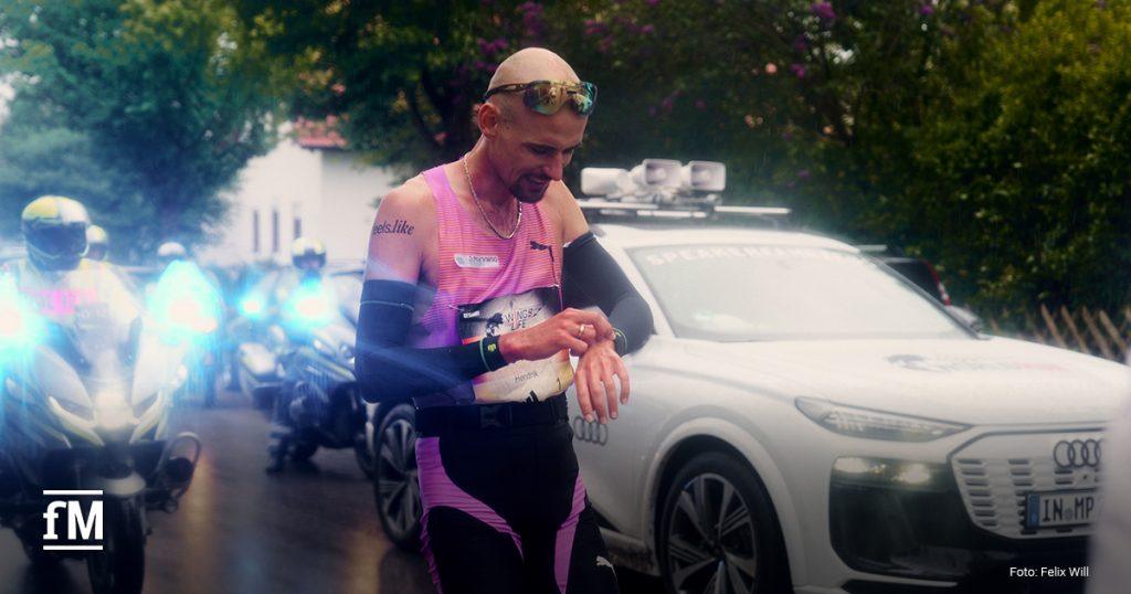 Ein männlicher Läufer (Hendrik Pfeiffer) im pinken feels.like-Outfit schaut während eines Rennens auf seine Uhr. Im Hintergrund begleiten Polizei-Motorräder und ein Audi-Fahrzeug mit der Aufschrift 'Speakercar' das Rennen.