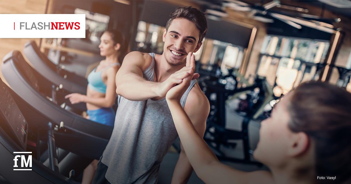 In einem modernen Fitnessstudio geben sich ein junger Mann und eine Frau, beide sportlich gekleidet, ein motivierendes High-Five. Der Mann trägt ein graues Tanktop und lächelt der Frau zu. Im Hintergrund trainieren weitere Personen auf Laufbändern, das Studio ist hell und großzügig mit vielen Fitnessgeräten ausgestattet. Links oben im Bild befindet sich der Schriftzug 'FLASHNEWS', das erste Wort ist grau, das zweite rot.