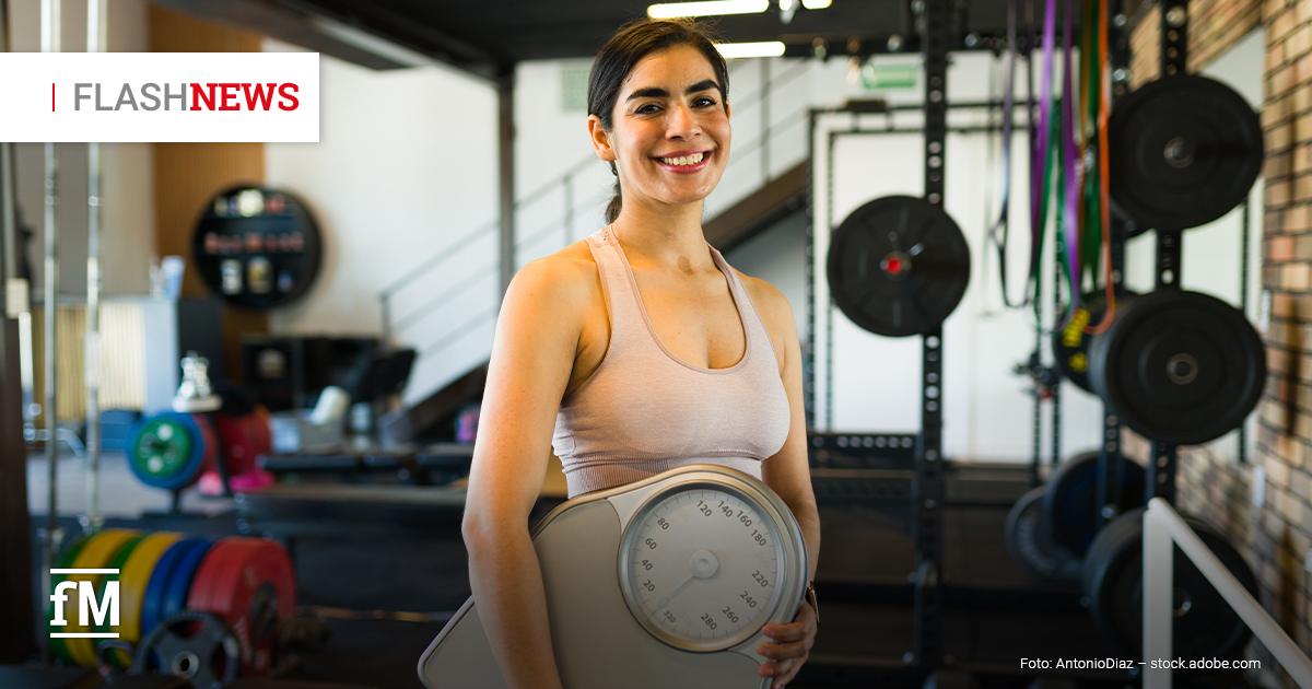 Eine junge Frau steht lächelnd in einem Fitnessstudio und hält eine große analoge Waage vor ihrem Oberkörper. Im Hintergrund sind Hanteln, Gewichtsscheiben und Sportgeräte sichtbar.
