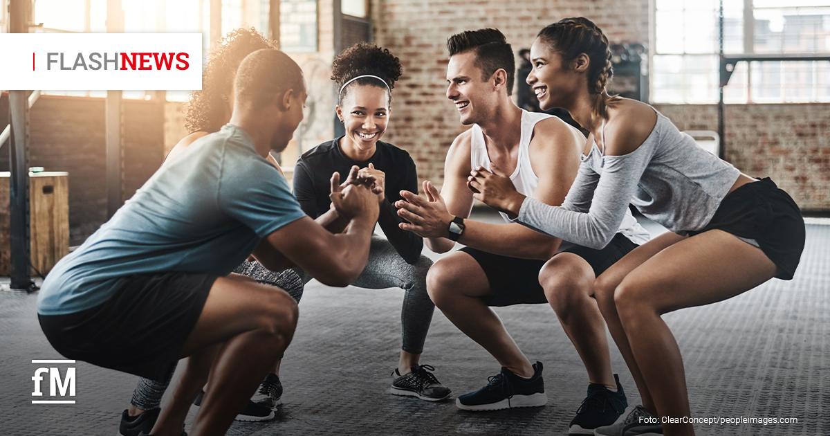 Fünf junge Erwachsene trainieren lachend in einem Fitnessstudio. Sie stehen sich paarweise gegenüber und machen gemeinsam Kniebeugen.