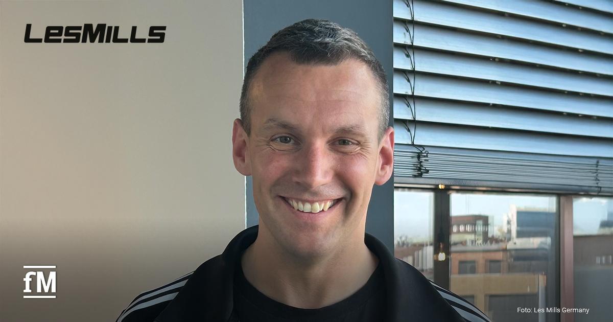 Jens Schulze, der neue CEO von Les Mills Germany, lächelt in die Kamera. Er trägt eine schwarzes Adidas-Trainingsjacke und Les Mills-T-Shirt und steht vor einem Fenster mit Jalousien. Im Hintergrund sind Gebäude und das Les Mills Logo zu sehen.