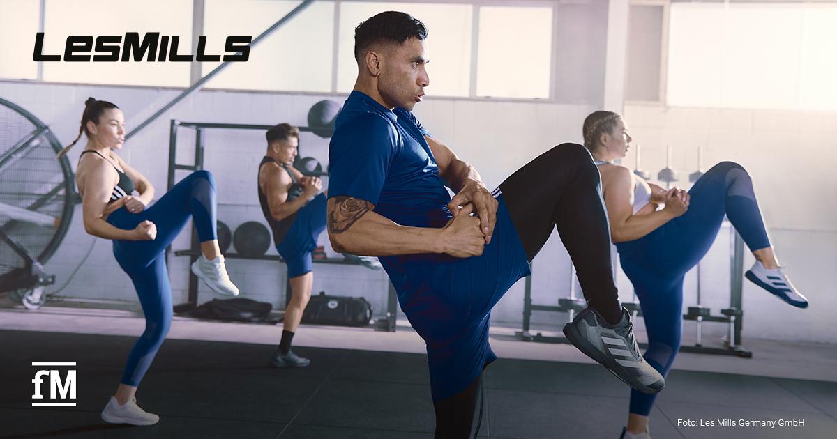 Fünf Personen führen eine dynamische Fitnessübung in einem Studio aus. Sie tragen Sportkleidung von 'Les Mills' und bewegen sich synchron. Der Fokus liegt auf einem Mann in blauer Kleidung.