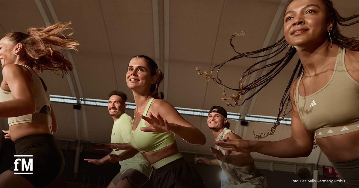 Gruppe junger Menschen tanzt und lacht bei einem energiegeladenen Workout. Alle tragen sportliche Kleidung in Beige und Grün. Die Stimmung ist motiviert und positiv. Markenlogo: 'Les Mills'