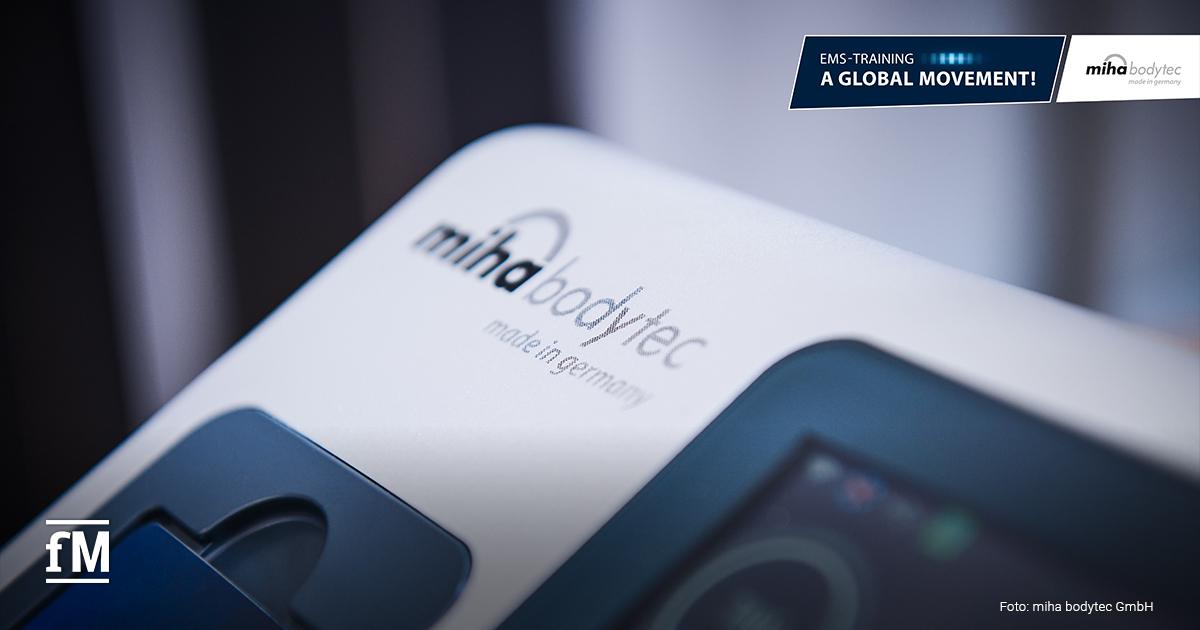 Nahaufnahme eines EMS-Geräts von 'miha bodytec'. Auf der Oberfläche steht 'made in Germany'. Oben rechts ist ein Banner mit dem Slogan 'A Global Movement!' zu sehen.