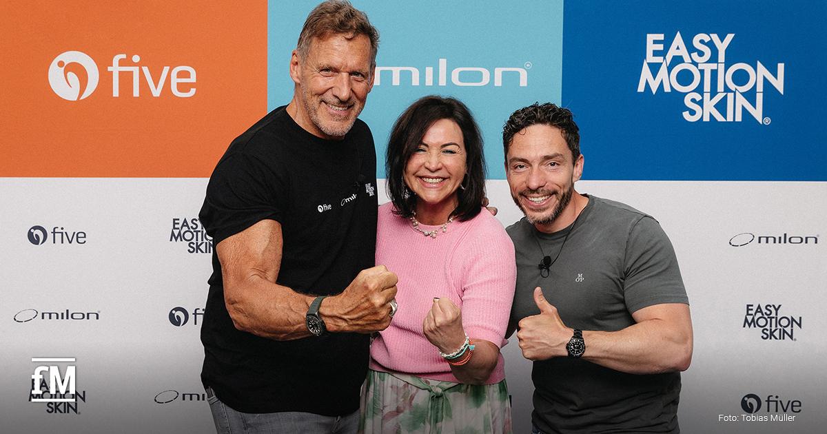 Drei Personen stehen lächelnd vor einer Sponsorenwand mit den Logos von five, milon und EasyMotionSkin. Links im Bild steht Ralf Moeller in schwarzem T-Shirt und Jeans, in der Mitte Sabine Beham in einem rosa Oberteil mit gemustertem Rock, rechts daneben Wolf Harwath in grauem T-Shirt und Jeans. Alle drei zeigen motiviert eine Faustpose in Richtung Kamera und strahlen Freude und Energie aus.