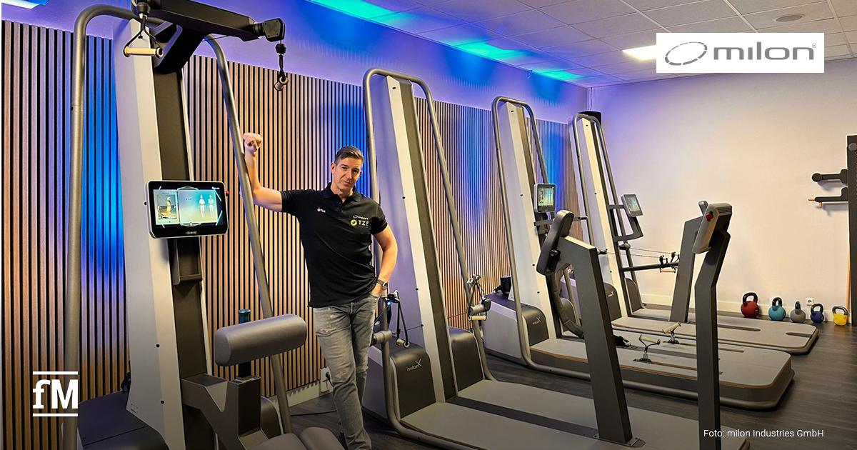 Dirk Rienesl steht in einem modernen Fitnessraum neben einem milon Trainingsgerät mit Bildschirm. Der Raum ist hell, mit Holzverkleidung an der Wand und blauem Licht. Im Hintergrund sind weitere Geräte und Kettlebells zu sehen.