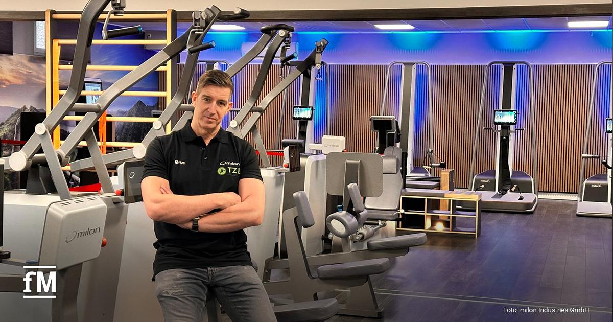 Dirk Rienesl steht mit verschränkten Armen vor modernen Trainingsgeräten im Fitnessstudio. Er trägt ein schwarzes Poloshirt, im Hintergrund leuchten Geräte in blauem Licht.