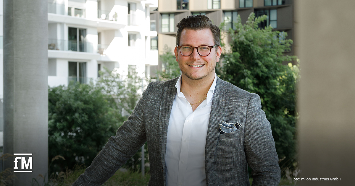 milon Industries GmbH Geschäftsführer Bernhard-Stefan Müller, ein Mann mit Brille im grauen Sakko und weißem Hemd, steht lächelnd im Freien vor moderner Wohnbebauung und grünen Bäumen.