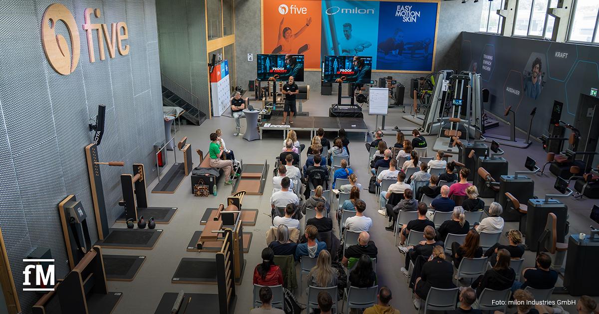 Teilnehmerinnen und Teilnehmer verfolgen eine Präsentation bei der milon & five Trainer Convention in einem modernen Trainingsraum mit Geräten und Bühne.