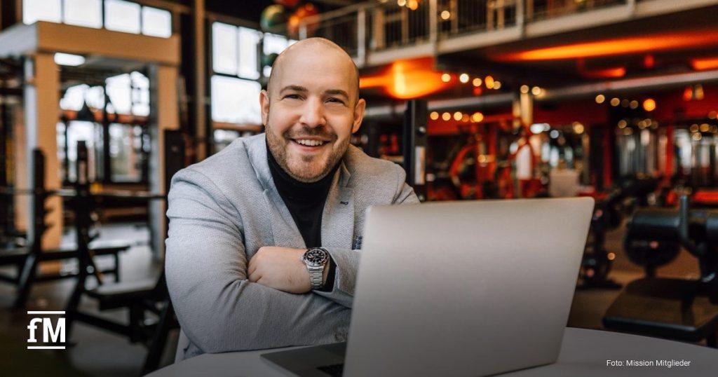 Ein Mann im grauen Sakko (Daniel Großgarten, Geschäftsführer von Mission Mitglieder) sitzt mit verschränkten Armen an einem Tisch vor einem geöffneten Laptop in einem modern ausgestatteten Fitnessstudio mit Trainingsgeräten im Hintergrund. Er lächelt freundlich in die Kamera.