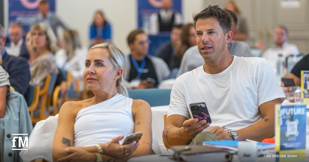 Zwei Teilnehmer eines Branchenevents sitzen aufmerksam im Publikum und verfolgen das Programm, beide halten ein Smartphone in der Hand.