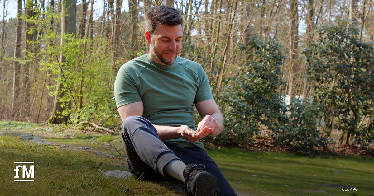 Ein Mann mit grünem T-Shirt sitzt im Schneidersitz auf einer Wiese im Wald und isst Pistazien; sein Blick ist auf die Hand gerichtet.