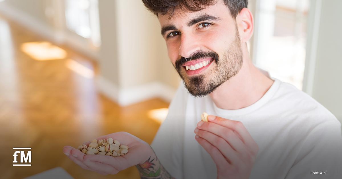 Ein junger Mann mit dunklem Bart, Nasenpiercing und Tattoos hält in einer Hand Pistazien und lacht, während er eine Pistazie zum Mund führt.