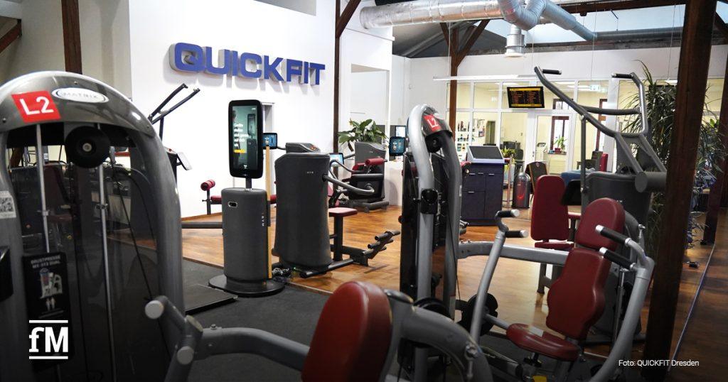 QUICKFIT-Bereich mit mehreren modernen Geräten. Im Hintergrund der Check-in-Bereich und eine Übersichtstafel. Die Gestaltung ist offen, mit Holz- und Glaselementen.