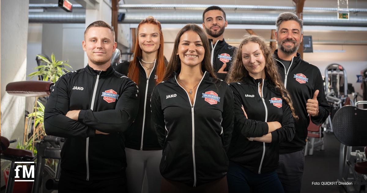 Sechs Mitarbeitende von QUICKFIT Dresden stehen im Fitnessstudio. Alle tragen schwarze JAKO-Jacken mit Logo. Sie lächeln motiviert in die Kamera und zeigen Teamgeist.