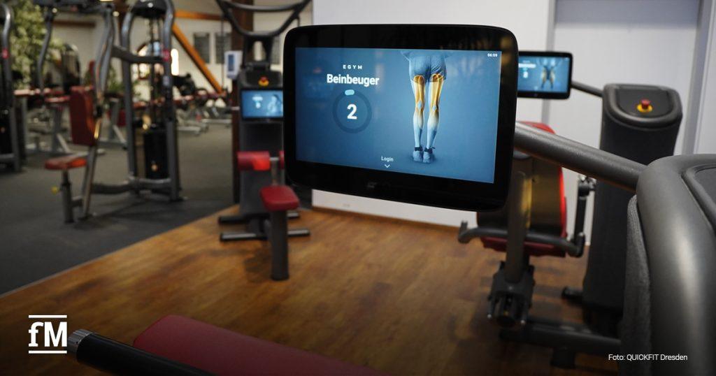 Nahaufnahme eines digitalen Trainingsgeräts für Beinbeuger im Studio. Der Bildschirm zeigt Muskelgruppen. Im Hintergrund sind weitere moderne Geräte sichtbar.