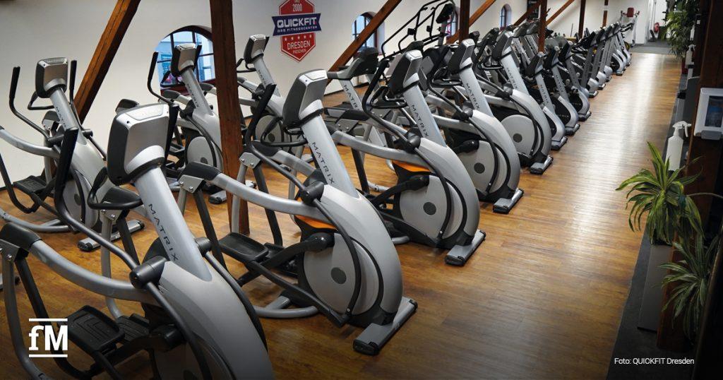Lange Reihe silberner Crosstrainer von MATRIX auf Holzfußboden. Der Trainingsbereich ist groß, ordentlich und lichtdurchflutet. Ein QUICKFIT-Wandlogo ist im Hintergrund sichtbar.