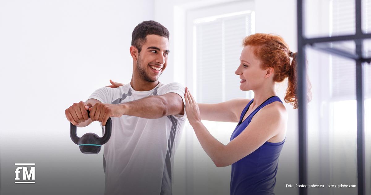 Eine Trainerin unterstützt einen jungen Mann beim Training mit einer Kettlebell – beide lächeln, die Atmosphäre ist hell und freundlich.