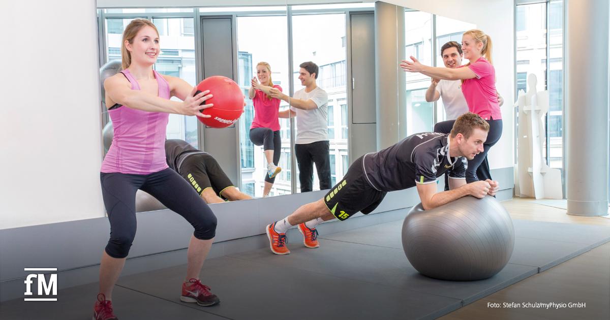 Mehrere Personen trainieren mit Medizinball und Gymnastikball in einem hellen Therapieraum mit Spiegelwand.