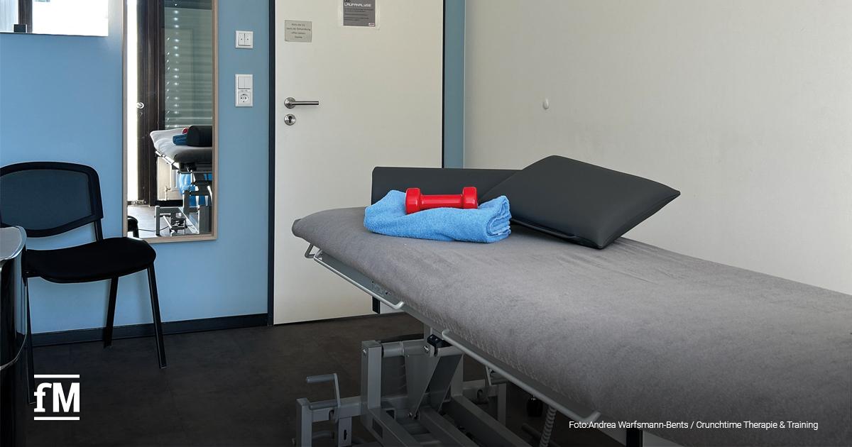 Therapieraum mit grauer Behandlungsliege, auf der ein gefaltetes blaues Handtuch und eine rote Kurzhantel liegen. Links stehen zwei schwarze Stühle, im Hintergrund eine weiße Tür und ein Spiegel, in dem der Raum mit Liege reflektiert wird.