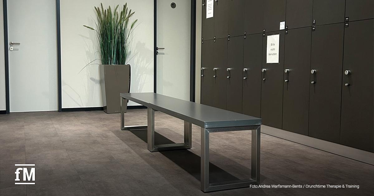Umkleideraum im Crunchtime mit dunklen Spinden an der Wand, mittig eine metallene Sitzbank auf grauem Boden. Links steht ein hoher Pflanzkübel mit Ziergras vor einer weißen Tür.