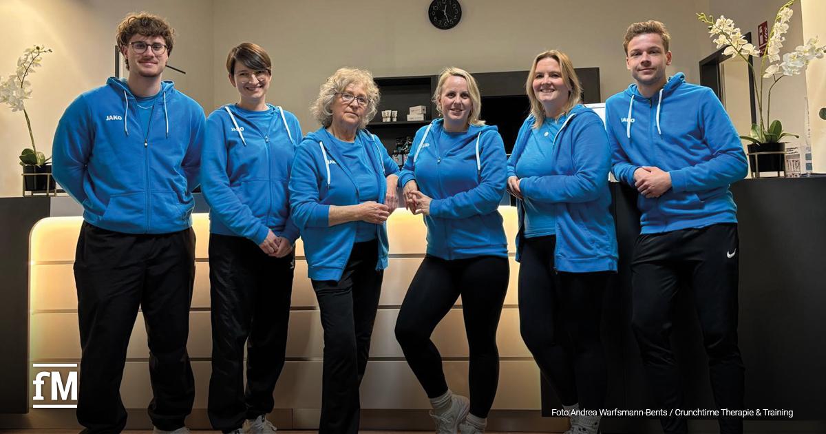 Sechs Mitarbeitende des Crunchtime Therapie & Training stehen nebeneinander vor dem beleuchteten Empfangstresen. Sie tragen blaue JAKO-Hoodies und schwarze Hosen, lächeln freundlich in die Kamera. Im Hintergrund sind dekorative Orchideen und eine Uhr an der Wand sichtbar.