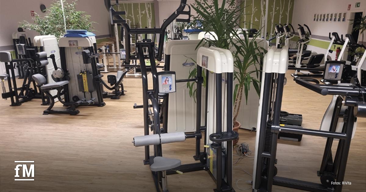Heller Trainingsraum mit modernen Kraftgeräten und Holzfußboden. Im Vordergrund mehrere Geräte mit integrierten Bildschirmen. Pflanzen lockern den Raum auf, im Hintergrund sind weitere Geräte sichtbar.