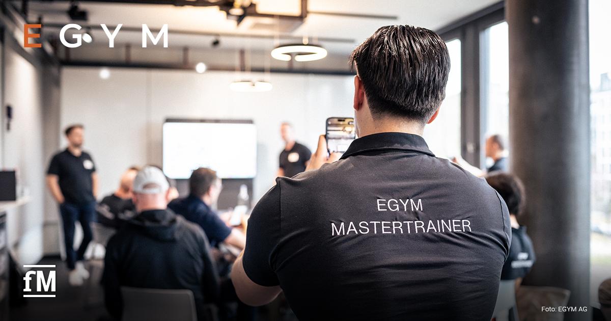 Eine Person mit 'EGYM MASTERTRAINER'-Shirt filmt eine Präsentation in einem modernen Schulungsraum mit mehreren Teilnehmenden und Vortragenden.