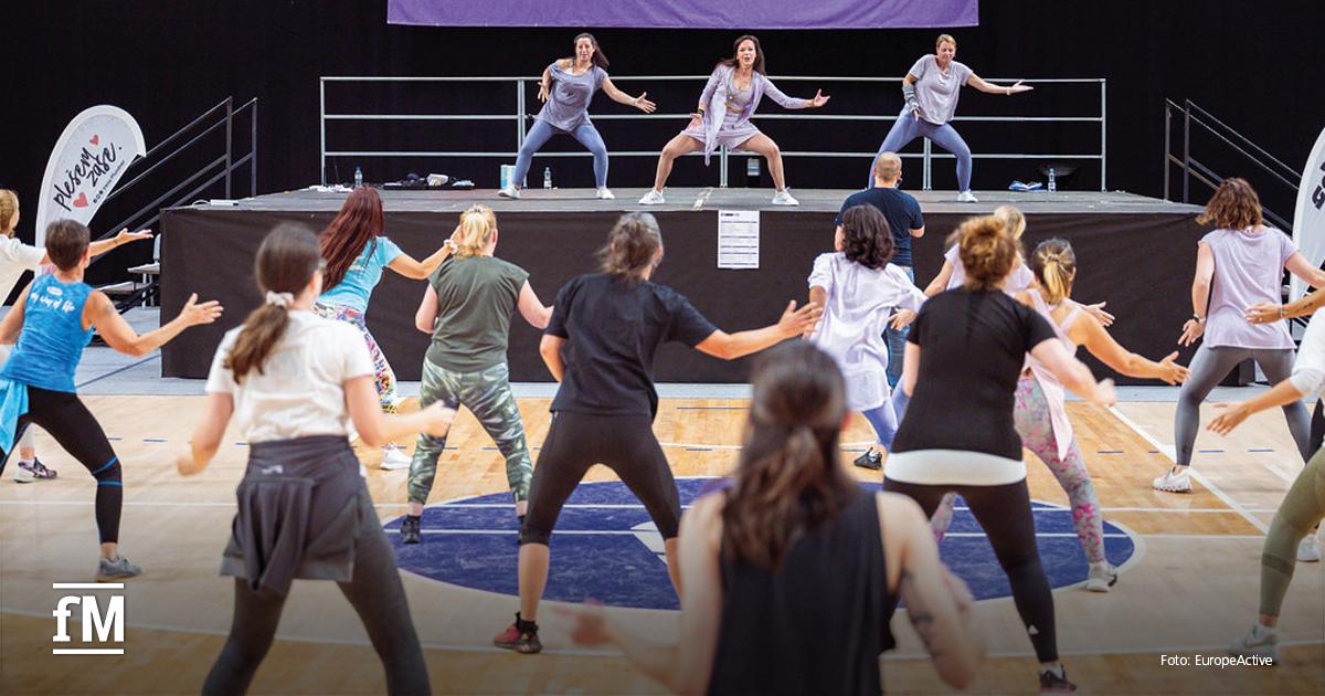 Gruppentraining mit etwa zwölf Teilnehmenden in einer Sporthalle; drei Trainerinnen auf einer Bühne geben rhythmische Bewegungen vor.