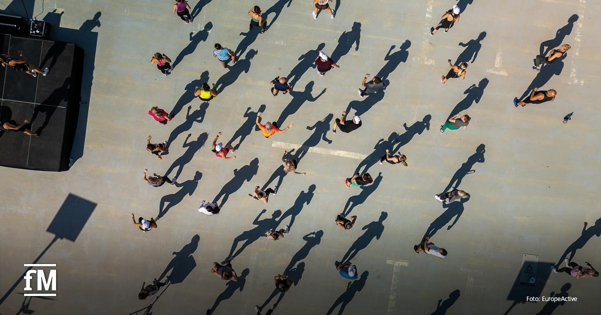 Drohnenaufnahme eines Outdoor-Fitnesskurses auf Betonboden; Teilnehmende stehen in Reihen mit langen Schatten links oben ein Podest mit Trainerinnen.