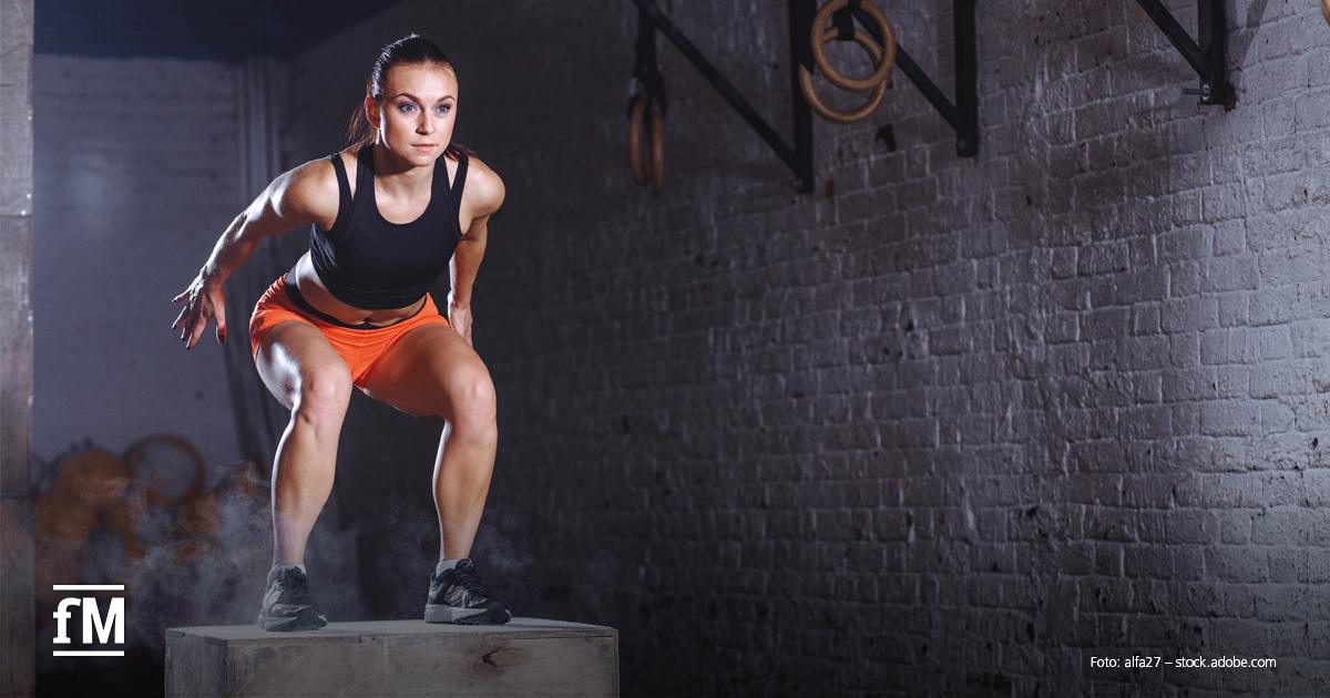 Eine sportliche Frau in Fitnesskleidung springt konzentriert auf eine Holzbox in einem Trainingsraum mit Ziegelwand.