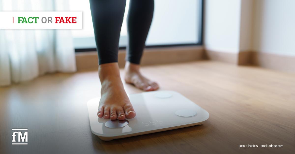 Barfüßige Person steht mit einem Fuß auf einer weißen digitalen Personenwaage auf Holzboden, der zweite Fuß befindet sich dahinter auf dem Boden