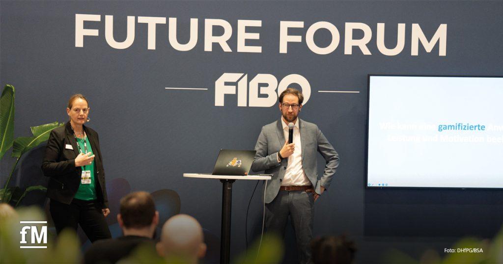 Zwei Vortragende stehen auf einer Bühne des Future Forum der FIBO. Links eine Frau mit Headset, rechts ein Mann im Anzug mit Mikrofon. Zwischen ihnen ein Stehtisch mit Laptop. Im Hintergrund ist ein großer Bildschirm zu sehen mit teilweise lesbarem Vortragstitel. Auf der Wand steht groß „FUTURE FORUM FIBO“. Vor der Bühne sitzt Publikum.