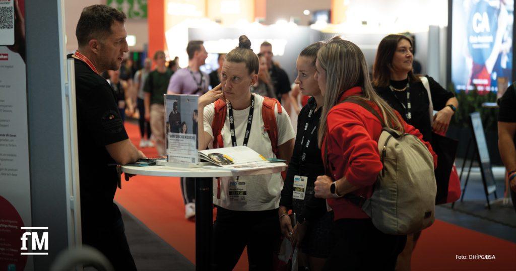 Mehrere Personen stehen an einem Messestand im Gespräch. Eine Frau in weißem T-Shirt hört aufmerksam zu, während ein Mann in schwarzem T-Shirt mit ihr spricht. Es liegt Infomaterial auf einem Stehtisch mit einem Aufsteller, auf dem „WIR SUCHEN DICH“ steht. Im Hintergrund sind weitere Messebesucher unterwegs.