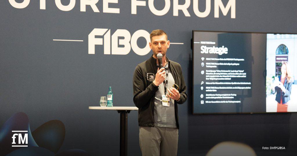 Ein Mann hält einen Vortrag auf dem „FIBO Future Forum“. Er spricht in ein Mikrofon und trägt eine schwarze Jacke mit Eventausweis. Neben ihm ein Stehtisch mit Wasserflasche. Hinter ihm ist eine Präsentation mit dem Titel „Strategie“ auf einem Bildschirm zu sehen.