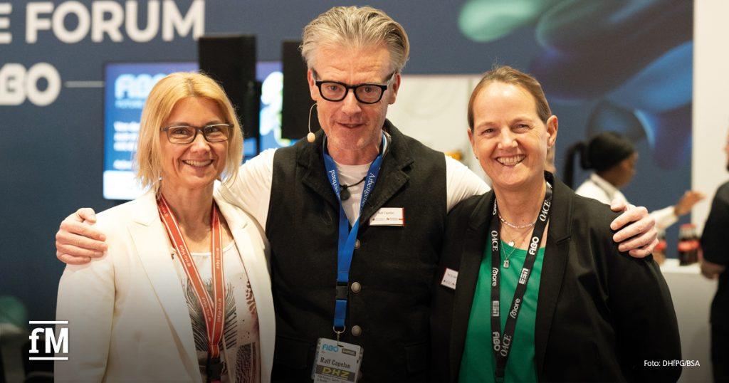 Drei Personen stehen Arm in Arm und lächeln in die Kamera. Links eine Frau mit blondem Haar und Brille im weißen Blazer, in der Mitte ein Mann mit grauem Haar und Brille im schwarzen Outfit, rechts eine Frau mit dunklem Blazer. Im Hintergrund ist das „FIBO Future Forum“-Logo sichtbar.