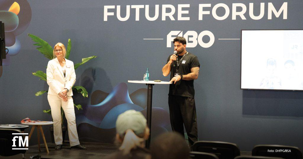Eine Frau im weißen Anzug steht auf der Bühne, neben ihr ein muskulöser Mann in schwarzem T-Shirt, der ins Mikrofon spricht. Beide stehen vor einem Hintergrund mit dem Logo „FUTURE FORUM FIBO“. Im Publikum sind mehrere Zuhörer zu sehen.
