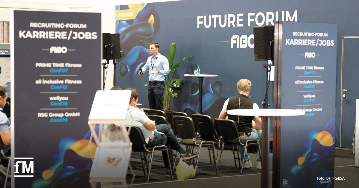 Ein Mann steht auf einer Bühne vor einem 'Future Forum'-Hintergrund auf der FIBO und spricht zu einem Publikum. Im Vordergrund stehen zwei Aufsteller mit Informationen zum Recruiting-Forum 'Karriere/Jobs' mit Unternehmenslogos.
