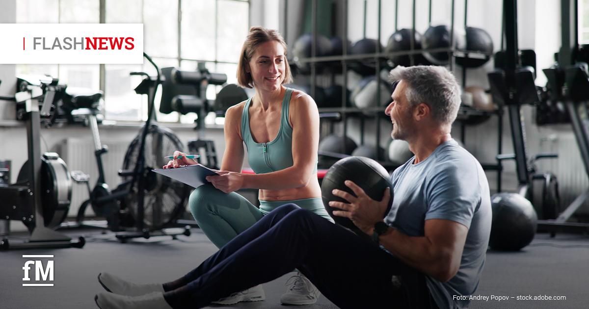 Eine Frau in türkisfarbener Sportkleidung kniet mit Klemmbrett vor einem Mann, der im Fitnessstudio mit einem Medizinball trainiert. Beide lächeln einander an.