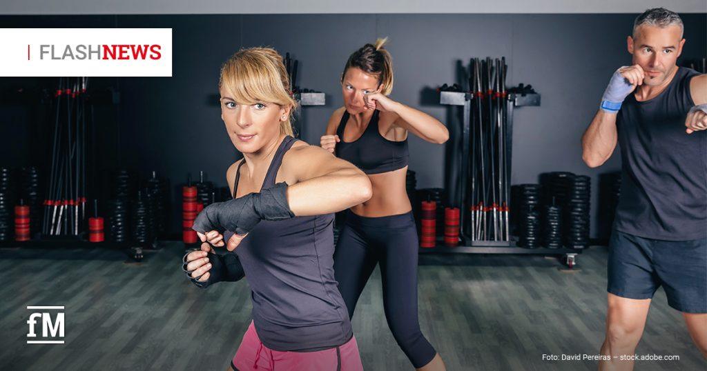 Drei Personen in Sportkleidung trainieren in einem Fitnessstudio gemeinsam eine Kampfsportübung mit Schlagbewegungen vor gestapelten Hanteln und Stange.