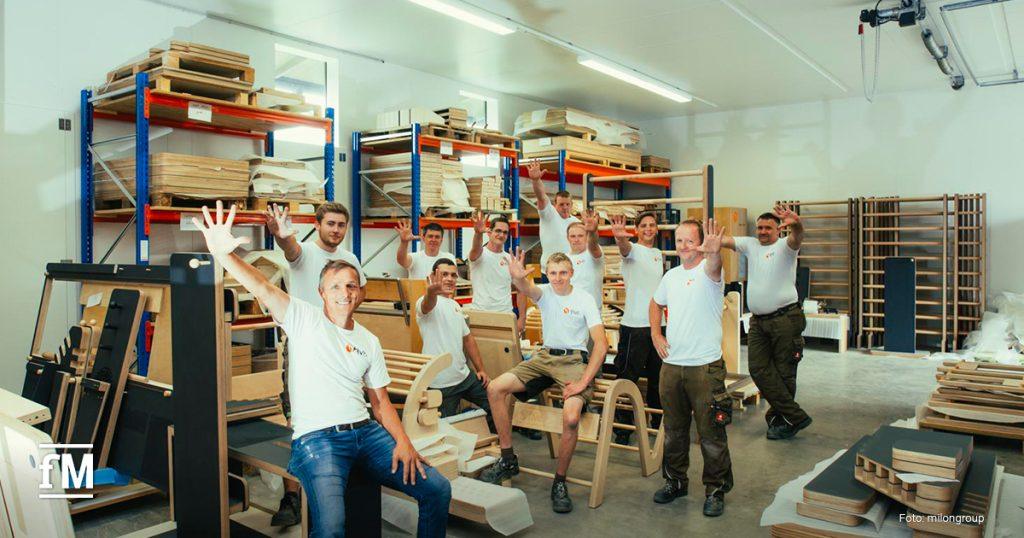 Produktionshalle von five mit einem Team aus Mitarbeitenden in weißem five-Shirt, die lächelnd in die Kamera winken, im Hintergrund Regale mit Holzmaterialien.