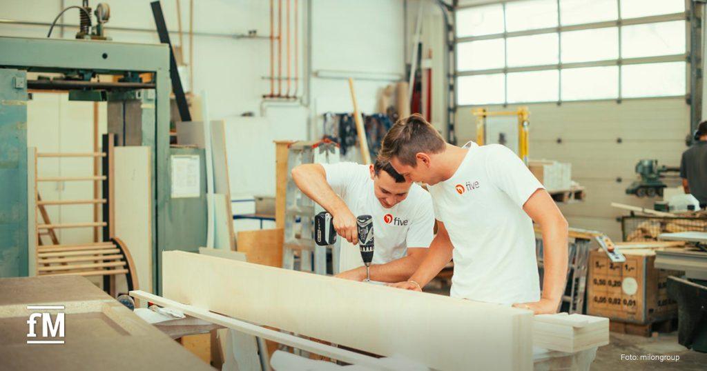 Zwei five-Mitarbeiter in weißem Shirt arbeiten gemeinsam an einem großen Holzbauteil in einer Werkstatt.