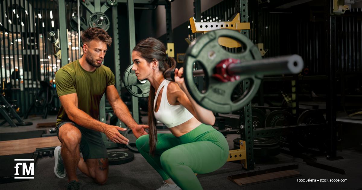 In einem modernen Fitnessstudio führt eine junge Frau in Sportkleidung eine Kniebeuge mit einer Langhantel auf dem Rücken aus. Ein Trainer in grünem T-Shirt und Shorts kniet vor ihr und gibt ihr konzentriert Anweisungen zur richtigen Technik. Im Hintergrund sind Hanteln, Spiegel und Fitnessgeräte zu sehen. Unten links befindet sich das fM-Logo.