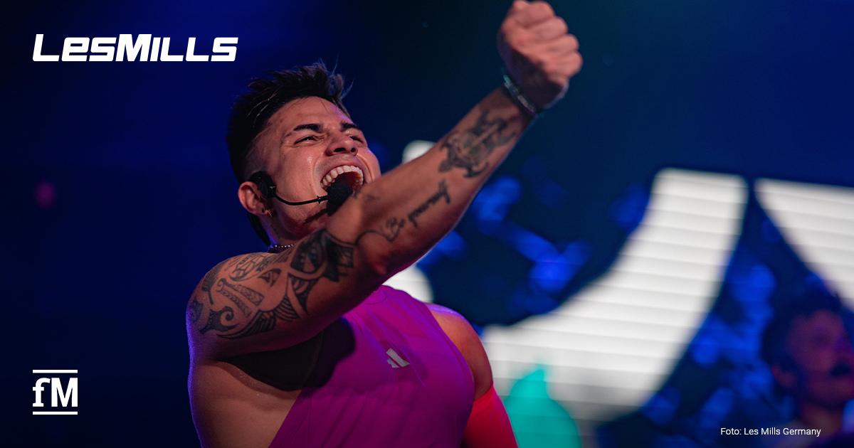 Les Mills Instruktor Sebastian Jaramillo mit auffälligen Tattoos, lila Muskelshirt und Headset ruft laut in Richtung Publikum, das nicht zu sehen ist, und reckt die Faust nach vorne; bunte Bühnenlichter im Hintergrund erzeugen eine kraftvolle Szenerie, während sein Gesichtsausdruck Leidenschaft und Motivation vermittelt.
