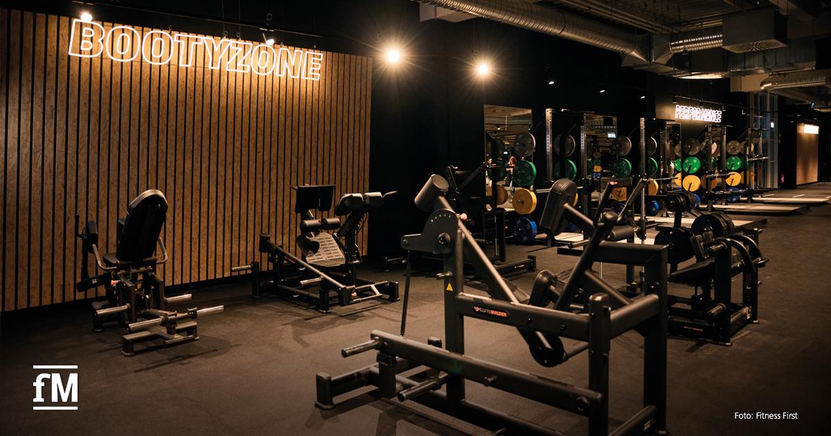 Dunkler Trainingsbereich mit Geräten für Gesäß- und Beintraining. An der Wand leuchtet der Schriftzug 'BOOTYZONE'. Im Hintergrund Hantelstationen und weitere Geräte mit bunten Gewichtsscheiben.