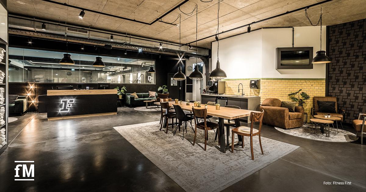 Moderner Lounge- und Empfangsbereich eines Fitnessstudios. Links Rezeption mit Logo, dahinter gemütliche Sitzgelegenheiten. Im Vordergrund steht ein großer Holztisch mit Stühlen, darüber hängen mehrere Lampen.