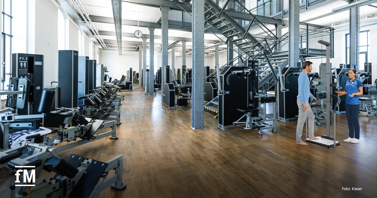 Helles, großzügiges Fitnessstudio mit modernen Geräten. Rechts steht ein Mann auf einer Analysewaage, betreut von einer Trainerin in blauer Kleidung. Industrielles Design mit Metallstützen und Treppe im Hintergrund.