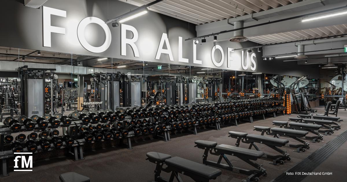 Eine modern ausgestattete Trainingsfläche mit zahlreichen Hanteln, Hantelbänken und Multifunktionsgeräten. An der spiegelverkleideten Wand leuchtet in großen Buchstaben der Slogan „FOR ALL OF US“.