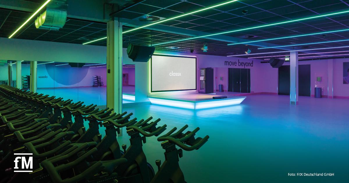 Ein großflächiger, futuristisch beleuchteter Kursraum mit Spinning-Rädern im Vordergrund. Auf der erhöhten Bühne in der Mitte steht „classx“, an der Wand dahinter der Claim „move beyond“.